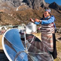 Kami Rita (Ang Nima's wife) heats water on a solar dish outside their home and Khunde Star Lodge faster than an electric kettle of the same size. Protector peak Khumbila rises above.